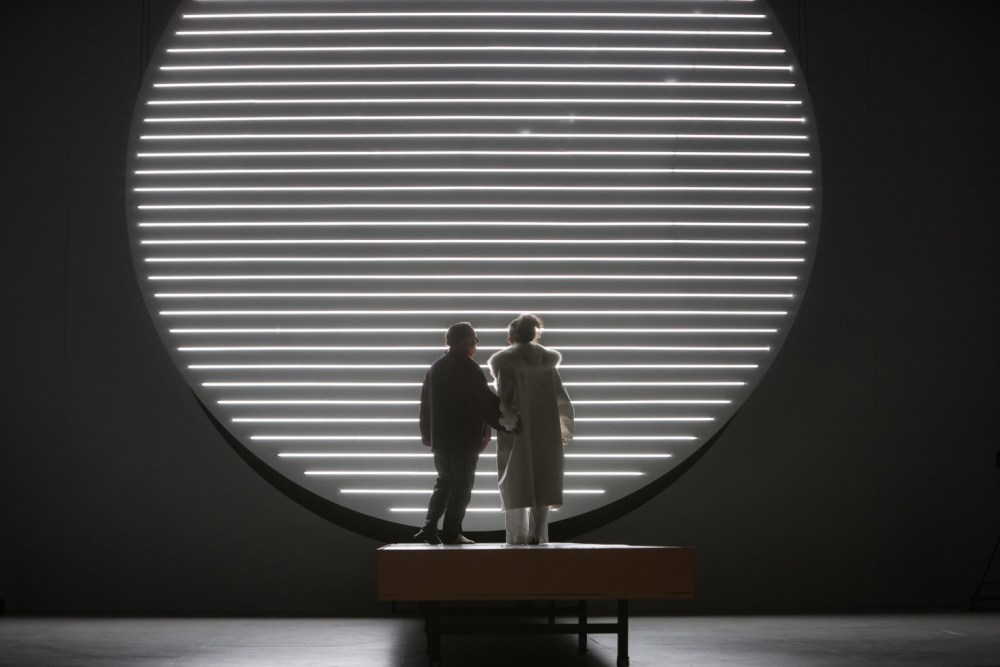 Two performers stand together on a stage, silhouetted against a large circular light installation with horizontal stripes. "Otello" 2025 at Theatre Bremen.