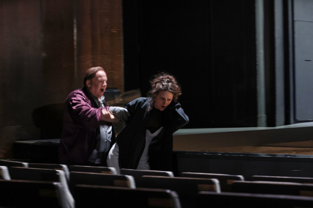 A dramatic scene from Verdi's "Otello" at Theater Bremen, featuring two characters in an intense moment. One figure, a man with short hair, passionately shouts while physically pulling a woman with curly hair towards him, conveying a sense of urgency and conflict in a theatrical setting.